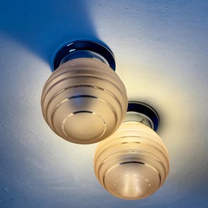May include: Two vintage ceiling lights with frosted glass globes and chrome accents. The globes have horizontal ribbed detailing and are mounted on a blue ceiling. The lights are illuminated, casting a warm glow.