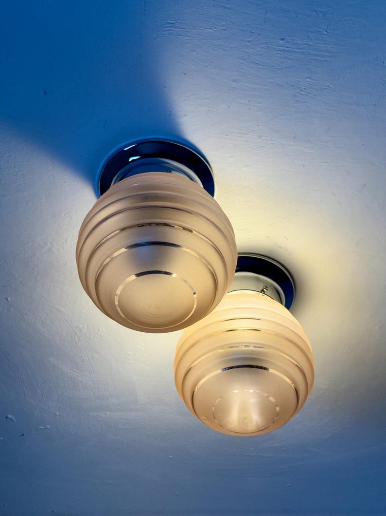 May include: Two vintage ceiling lights with frosted glass globes and chrome accents. The globes have horizontal ribbed detailing and are mounted on a blue ceiling. The lights are illuminated, casting a warm glow.