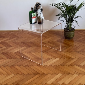 Puede incluir: Una mesa auxiliar transparente de acrílico con un diseño curvo. Sobre la mesa hay una botella verde con una etiqueta, una figura de perro de cerámica y un vaso marrón. Una planta en maceta está a un lado. La mesa está sobre un suelo de madera.