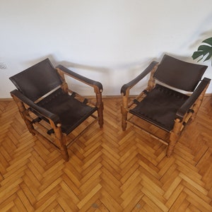 May include: Two brown leather chairs with wooden frames. The chairs are on a wooden floor with a herringbone pattern.