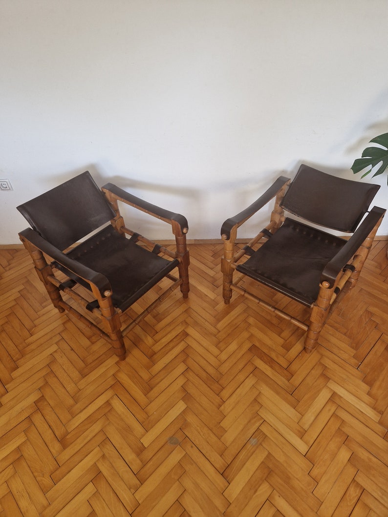 May include: Two brown leather chairs with wooden frames. The chairs are on a wooden floor with a herringbone pattern.