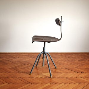 Puede incluir: Una silla giratoria de estilo industrial vintage con estructura de metal y asiento de madera marr&oacute;n oscuro. La silla tiene un dise&ntilde;o &uacute;nico con un respaldo curvo y una base de cuatro patas.