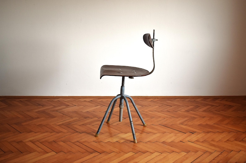 Puede incluir: Una silla giratoria de estilo industrial vintage con estructura de metal y asiento de madera marr&oacute;n oscuro. La silla tiene un dise&ntilde;o &uacute;nico con un respaldo curvo y una base de cuatro patas.