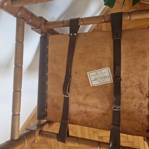May include: A wooden chair with a brown leather seat and back. The chair has two brown leather straps that are attached to the back of the chair. The straps have metal buckles. The chair has a label on the seat that says "MORRIS VOGELMAN, FABRICANTE DE SILLAS, BUENOS AIRES".
