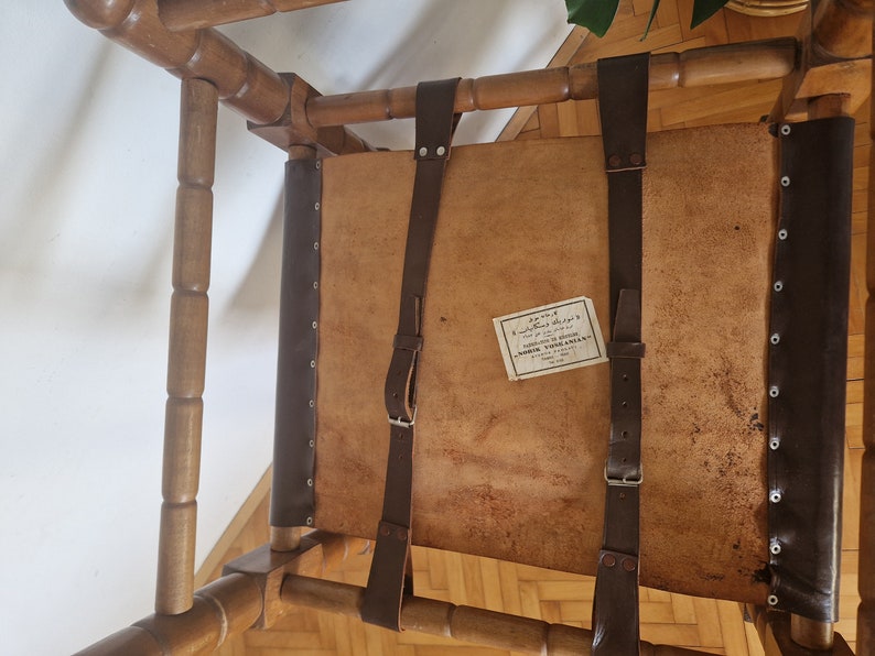 May include: A wooden chair with a brown leather seat and back. The chair has two brown leather straps that are attached to the back of the chair. The straps have metal buckles. The chair has a label on the seat that says "MORRIS VOGELMAN, FABRICANTE DE SILLAS, BUENOS AIRES".