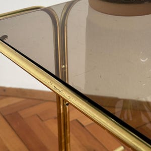 May include: A gold metal serving cart with a glass top. The cart has a simple design with a rectangular top and four legs. The cart is on a wooden floor.