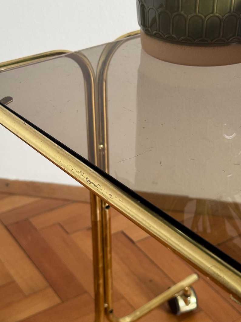May include: A gold metal serving cart with a glass top. The cart has a simple design with a rectangular top and four legs. The cart is on a wooden floor.