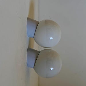 May include: Two white globe-shaped wall sconces with a light blue base are mounted on a light-colored wall. The sconces have a smooth, glossy finish, and the light blue base provides a subtle contrast.
