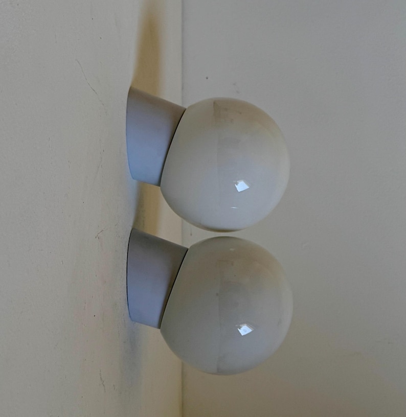 May include: Two white globe-shaped wall sconces with a light blue base are mounted on a light-colored wall. The sconces have a smooth, glossy finish, and the light blue base provides a subtle contrast.