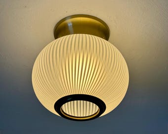 May include: A round, cream-colored ceiling light fixture with vertical ribbed detailing. The light has a brass-colored base and a black ring around the bottom. The light is illuminated, casting a warm glow.