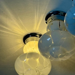 May include: Close-up of a modern ceiling light fixture with multiple white, orb-shaped glass shades. The shades have a textured, speckled pattern. The fixture has chrome accents and is mounted on a white ceiling. The light casts a warm glow.