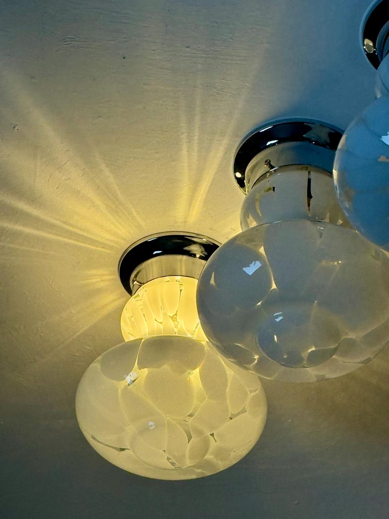May include: Close-up of a modern ceiling light fixture with multiple white, orb-shaped glass shades. The shades have a textured, speckled pattern. The fixture has chrome accents and is mounted on a white ceiling. The light casts a warm glow.