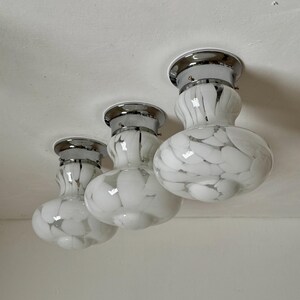 May include: Three white and gray speckled glass ceiling lights with chrome trim. The lights have a rounded, bulbous shape and are mounted on a white ceiling. The glass has a speckled pattern, creating a unique design.