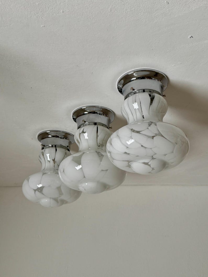 May include: Three white and gray speckled glass ceiling lights with chrome trim. The lights have a rounded, bulbous shape and are mounted on a white ceiling. The glass has a speckled pattern, creating a unique design.