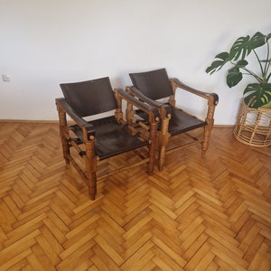 May include: Two brown leather chairs with wooden frames. The chairs have a simple design with a woven pattern on the back and arms. The chairs are sitting on a wooden floor with a herringbone pattern.