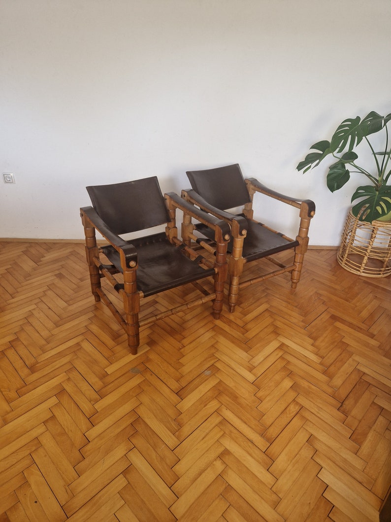 May include: Two brown leather chairs with wooden frames. The chairs have a simple design with a woven pattern on the back and arms. The chairs are sitting on a wooden floor with a herringbone pattern.