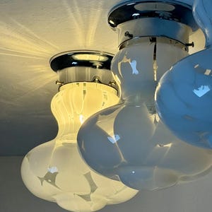 May include: Close-up of three white and chrome ceiling lights. Each light has a bulbous, rounded shape with a chrome base and a white glass shade. The lights are mounted on a white ceiling, with light reflecting off the surfaces.