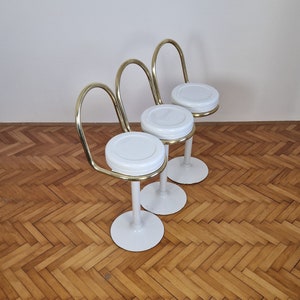 May include: Three white and gold bar stools with curved backs. The stools have a white round seat and a white base.