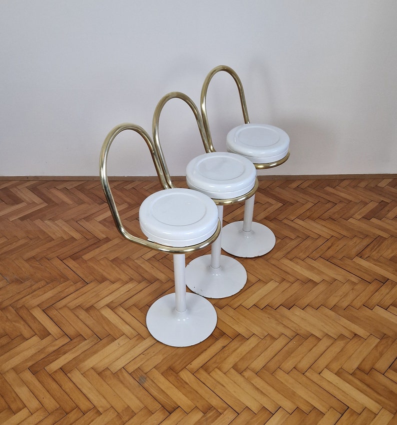 May include: Three white and gold bar stools with curved backs. The stools have a white round seat and a white base.
