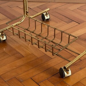 May include: A gold metal rolling stand with a wire basket underneath. The stand has four wheels and is on a wooden floor.