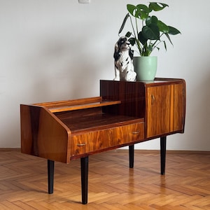 Puede incluir: Una mesa auxiliar de estilo moderno de mediados de siglo con un acabado en chapa de madera y patas negras. La mesa tiene un cajón, un estante y una puerta de armario. Una pequeña figurita de perro de cerámica y una planta en maceta están encima de la mesa.
