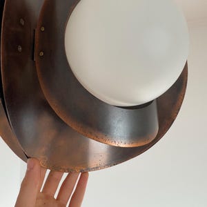 Puede incluir: Una l&aacute;mpara colgante de tono cobre con una pantalla de vidrio blanco. La l&aacute;mpara tiene un dise&ntilde;o en capas y org&aacute;nico.