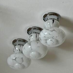 May include: Three flush-mount ceiling lights with a chrome finish and white glass globes. The globes have a speckled pattern of black and white. The lights are mounted on a white ceiling.