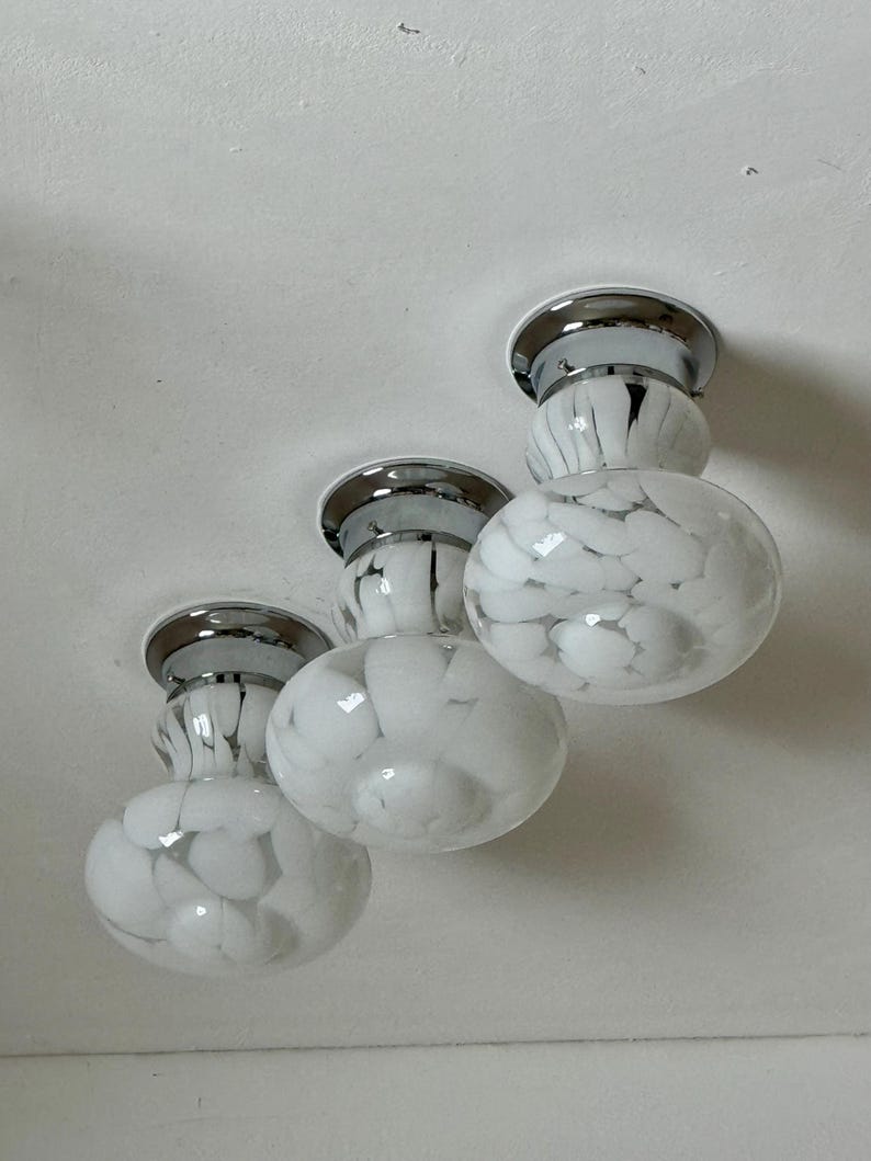 May include: Three flush-mount ceiling lights with a chrome finish and white glass globes. The globes have a speckled pattern of black and white. The lights are mounted on a white ceiling.