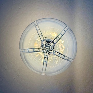 May include: A circular light fitting, viewed from above, with a star-shaped metal frame. The glass has a frosted appearance, and the fixture emits a warm light. The metal frame is silver, with small decorative rivets.