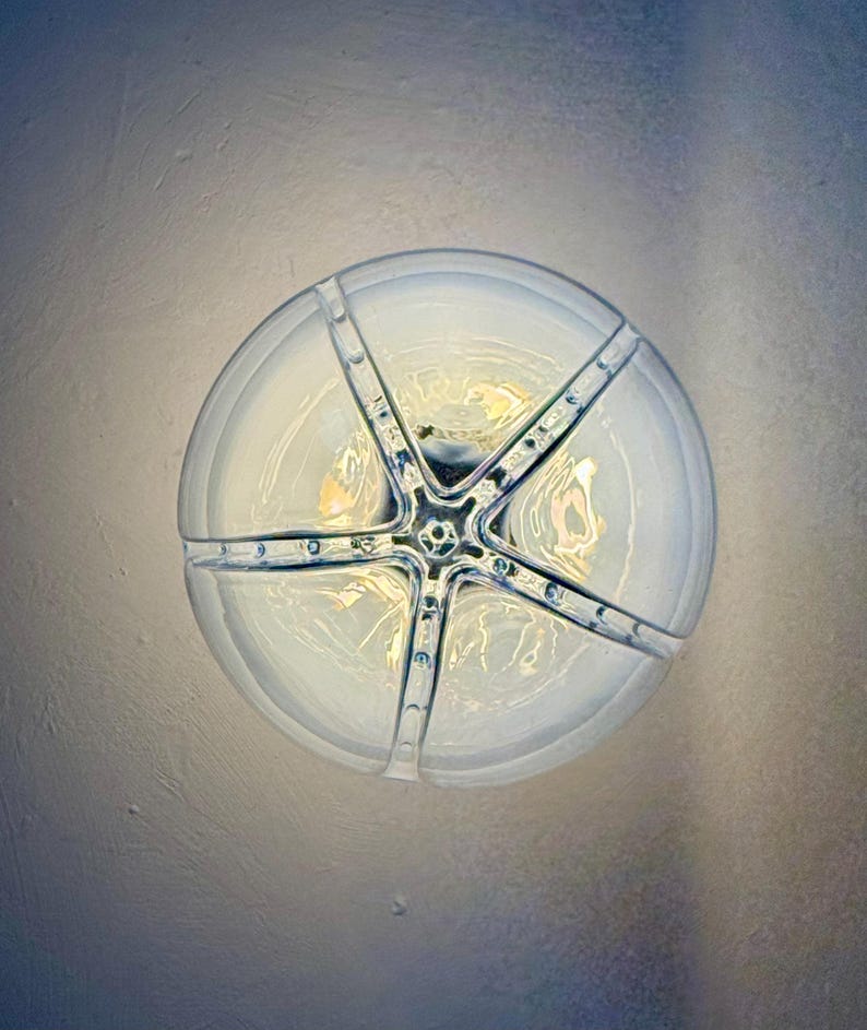 May include: A circular light fitting, viewed from above, with a star-shaped metal frame. The glass has a frosted appearance, and the fixture emits a warm light. The metal frame is silver, with small decorative rivets.