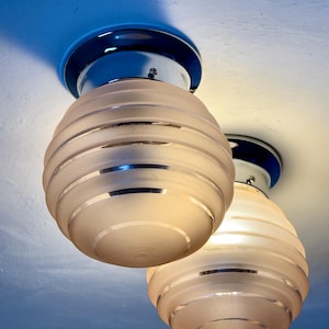 May include: Two Art Deco-style ceiling light fixtures. Each fixture has a frosted glass globe with horizontal bands, a chrome base, and a dark blue circular mounting plate. The lights are mounted on a white ceiling.
