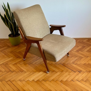 May include: A mid-century modern armchair with a beige fabric upholstery and wooden arms and legs. The chair is on a wooden floor with a herringbone pattern.