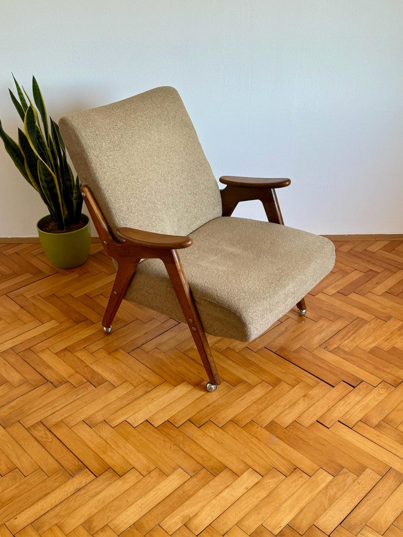May include: A mid-century modern armchair with a beige fabric upholstery and wooden arms and legs. The chair is on a wooden floor with a herringbone pattern.