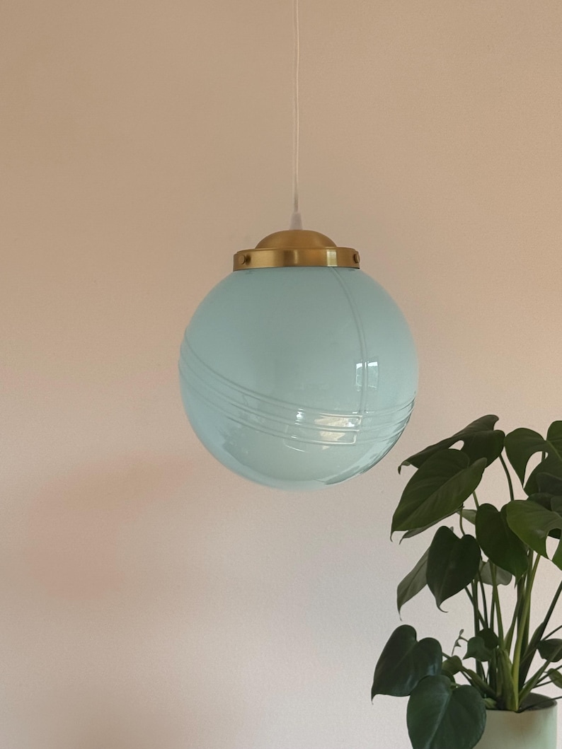 May include: A light blue glass globe pendant light with a gold metal fixture. The light is hanging from a white cord.