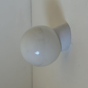 May include: A white, spherical wall-mounted light fixture. The light has a round, bulbous shape and is attached to the wall with a short, cylindrical arm. The fixture is made of a matte white material.