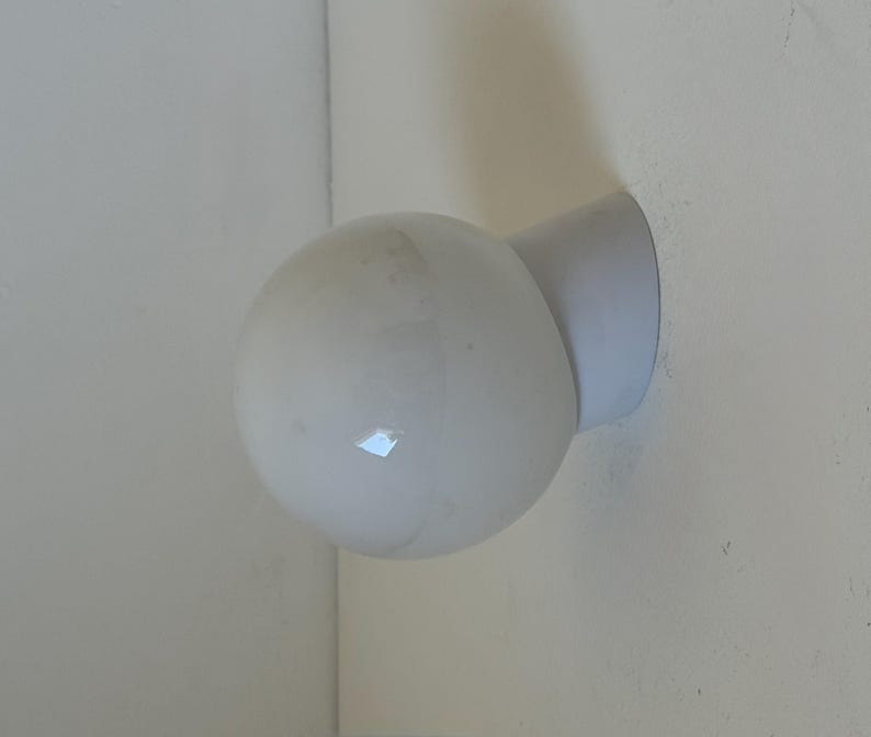 May include: A white, spherical wall-mounted light fixture. The light has a round, bulbous shape and is attached to the wall with a short, cylindrical arm. The fixture is made of a matte white material.