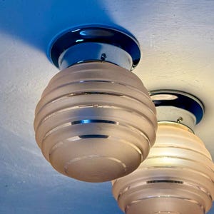 May include: Two vintage ceiling light fixtures. Each light has a frosted glass shade with horizontal bands and a chrome base. The shades are a soft pink color, and the bases are a dark blue. The lights are mounted on a white ceiling.