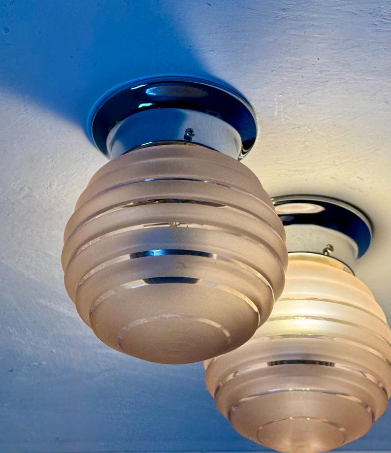 May include: Two vintage ceiling light fixtures. Each light has a frosted glass shade with horizontal bands and a chrome base. The shades are a soft pink color, and the bases are a dark blue. The lights are mounted on a white ceiling.