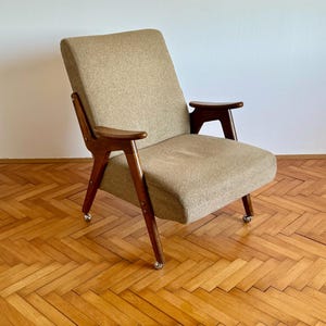 May include: A mid-century modern armchair with a beige fabric upholstery and wooden legs and arms. The chair is on a wooden floor with a herringbone pattern.