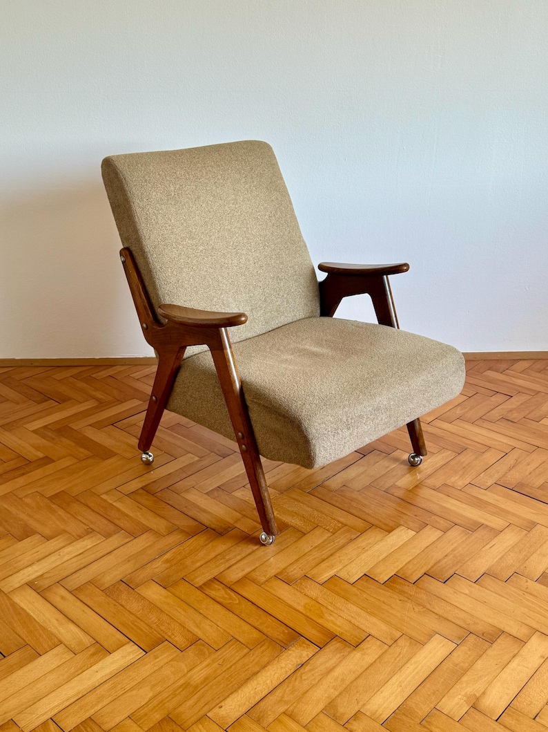 May include: A mid-century modern armchair with a beige fabric upholstery and wooden legs and arms. The chair is on a wooden floor with a herringbone pattern.