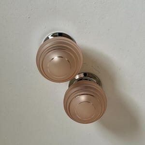 May include: Two matching, round door knobs. The knobs are a soft, muted pink color with a frosted glass appearance. Each knob has a series of concentric rings and a polished chrome base. The knobs are mounted on a white wall.