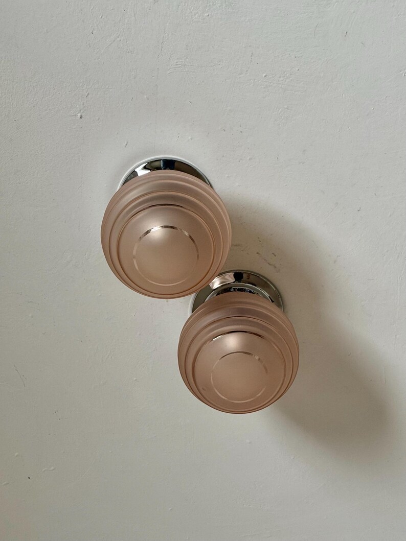 May include: Two matching, round door knobs. The knobs are a soft, muted pink color with a frosted glass appearance. Each knob has a series of concentric rings and a polished chrome base. The knobs are mounted on a white wall.
