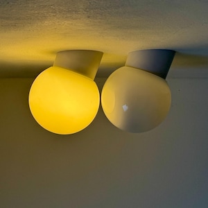 May include: Two modern, spherical ceiling lights. One is illuminated with a warm yellow glow, the other is off, appearing white. Each light is connected to a gray, angled base, mounted on a neutral-toned ceiling.