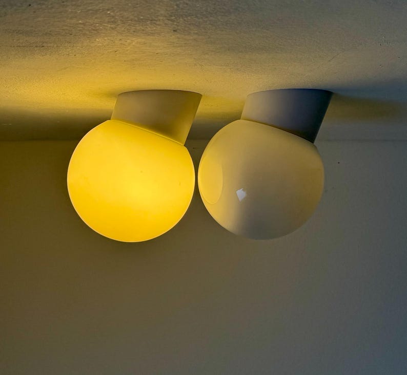 May include: Two modern, spherical ceiling lights. One is illuminated with a warm yellow glow, the other is off, appearing white. Each light is connected to a gray, angled base, mounted on a neutral-toned ceiling.