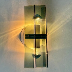 May include: A contemporary wall sconce with a rectangular, grey-tinted glass shade. The sconce has a vertical design with a light bulb in the centre, and is mounted on a white wall. The light is on, casting a warm glow.