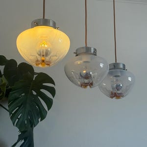 Op de afbeelding: Drie hanglampen met matglazen kappen en zichtbare gloeilampen hangen aan bruine snoeren. De kappen hebben een decoratief ontwerp en zijn bevestigd aan zilverkleurige armaturen. Een grote groene plant is zichtbaar.