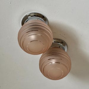 May include: Two ceiling-mounted light fixtures. Each fixture has a circular chrome base and a frosted, peach-colored glass shade. The shades are ribbed with a rounded, bulbous shape. The fixtures are mounted on a white ceiling.