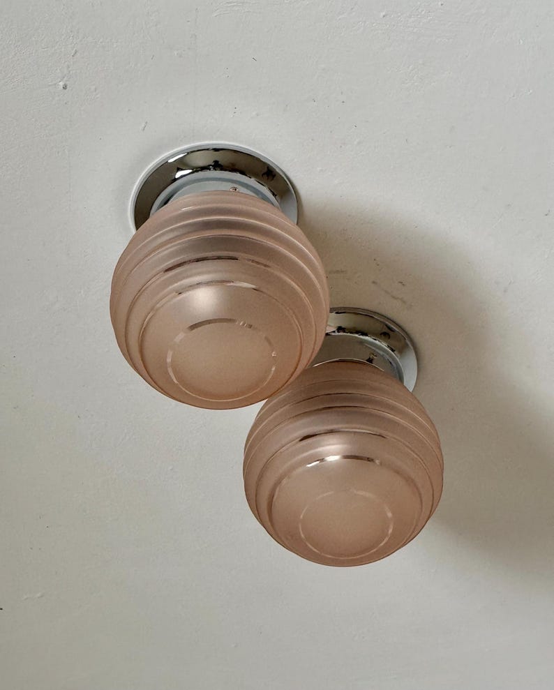 May include: Two ceiling-mounted light fixtures. Each fixture has a circular chrome base and a frosted, peach-colored glass shade. The shades are ribbed with a rounded, bulbous shape. The fixtures are mounted on a white ceiling.