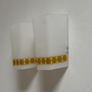 Puede incluir: Dos apliques de pared rectangulares blancos con un patrón geométrico amarillo cerca de la parte inferior. Los apliques están montados en una pared blanca, proporcionando una solución de iluminación sencilla y moderna.