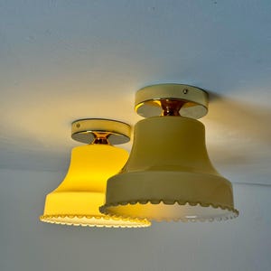 Pode incluir: Duas luminárias de teto vintage de vidro amarelo com borda ondulada. As luzes têm uma base de cor latão e são montadas em um teto branco. As abajures têm formato de sino e emitem um brilho quente.